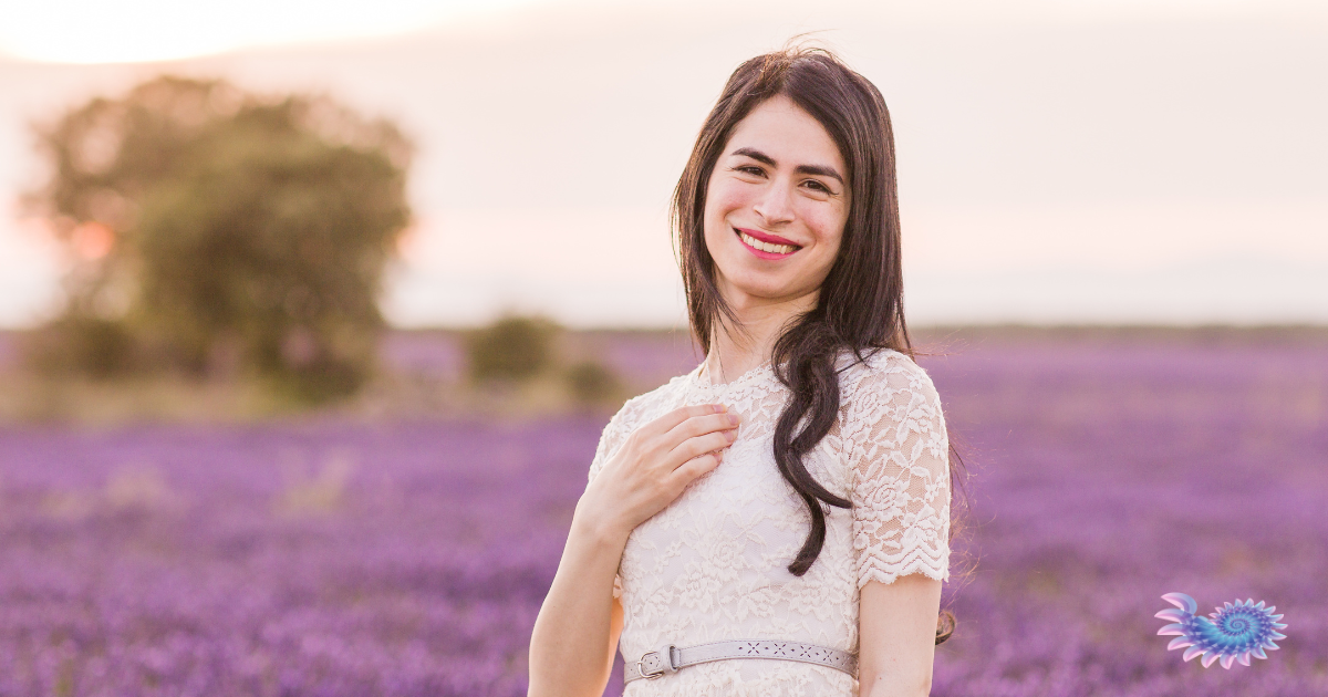 Beautiful transgender person wearing a romantic style dress standing in a field of lavender at sunset smiling beatifically at the viewer
