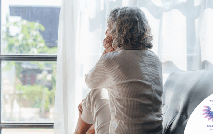Older lonely-seeming person staring out of the window from their couch, possibly contemplating their anxiety about how the world and society is perceiving their transgender child
