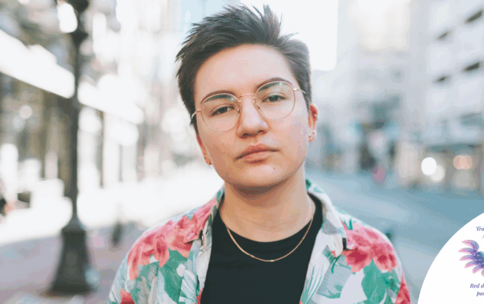 Trans masculine presenting person standing in a city, staring down the camera
